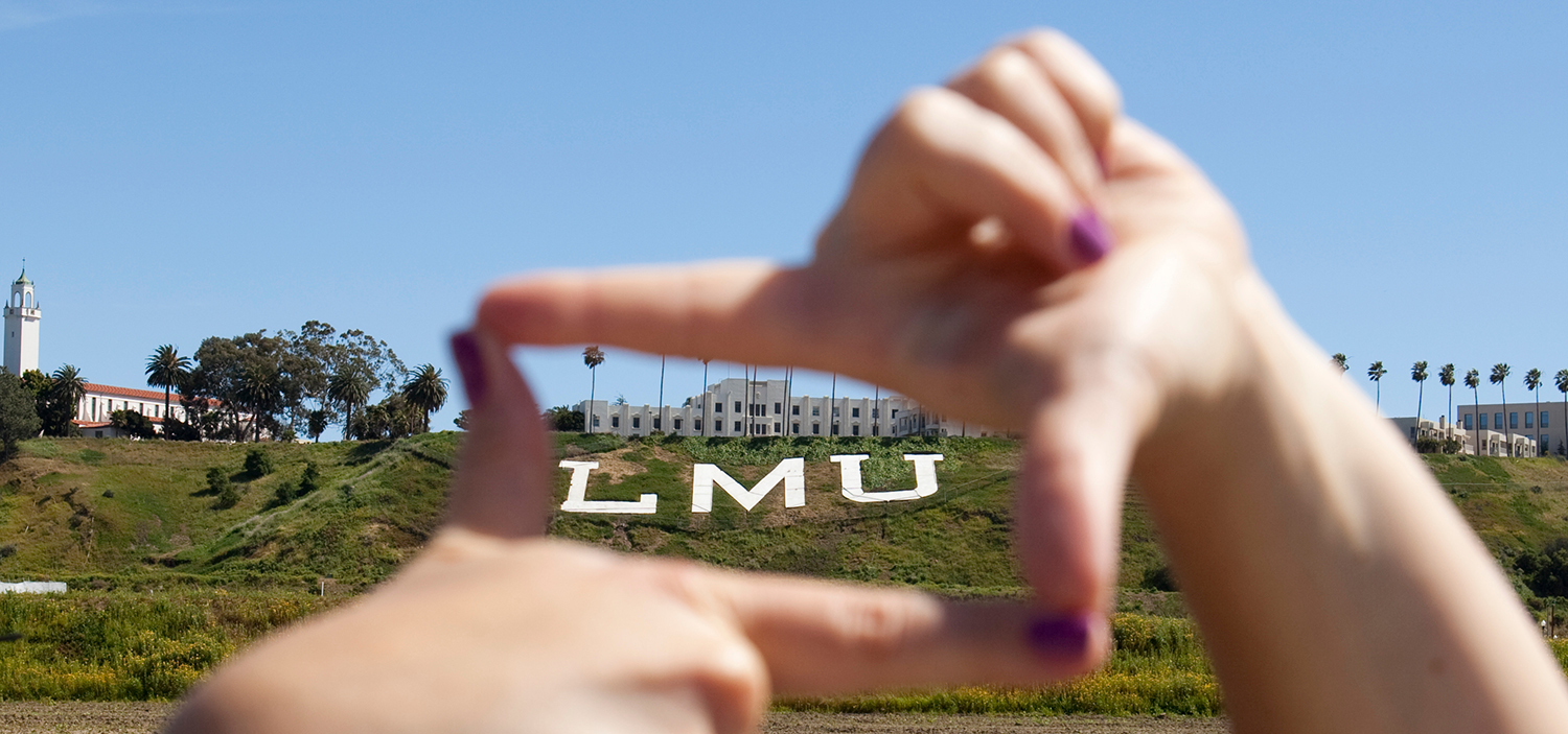 Large LMU letters placed on the side of the bluff with St. Robert's Hall on the top of the cliff, framed by a student's fingers like how a film director frames a shot.